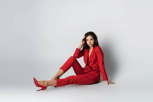 Pleine longueur de femme à la mode en costume rouge posant tout en étant assis sur le gris — Photo de stock