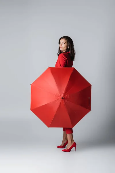 Longitud completa de la mujer morena en traje rojo de pie con paraguas en gris - foto de stock