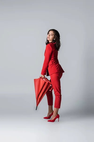 Full length of brunette woman in suit standing with red umbrella on grey — Stock Photo