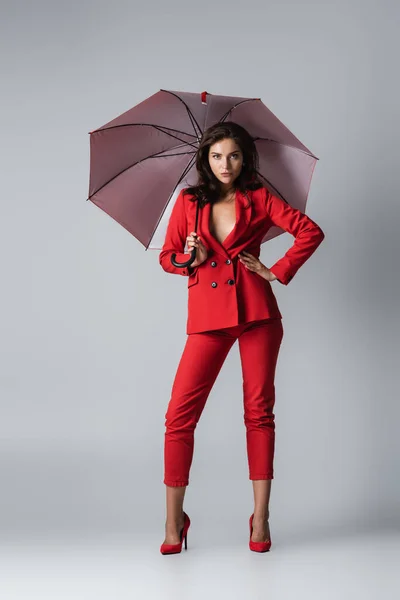 Full length of brunette woman in red suit standing with hand on hip under umbrella on grey — Stock Photo