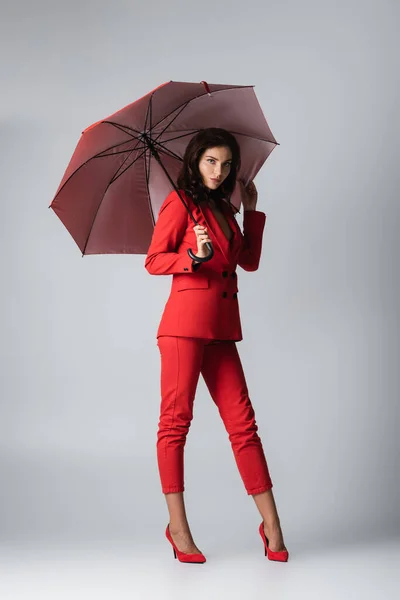 Full length of young woman in red suit standing under umbrella on grey — Stock Photo