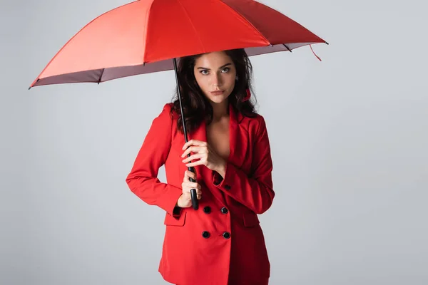 Брюнетка в красном костюме стоящая под зонтиком изолированная на сером — стоковое фото