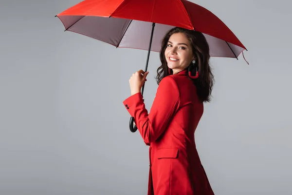 Felice giovane donna in abito rosso in piedi sotto l'ombrello isolato su grigio — Foto stock