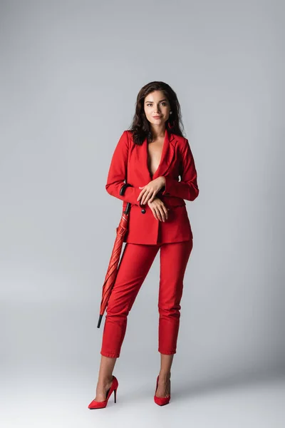 Pleine longueur de femme heureuse en costume rouge debout avec parapluie sur gris — Photo de stock