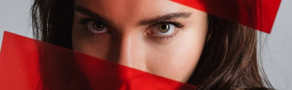 Обрезанный вид молодой женщины, смотрящей в камеру через красное стекло, изолированное на сером, баннер — стоковое фото