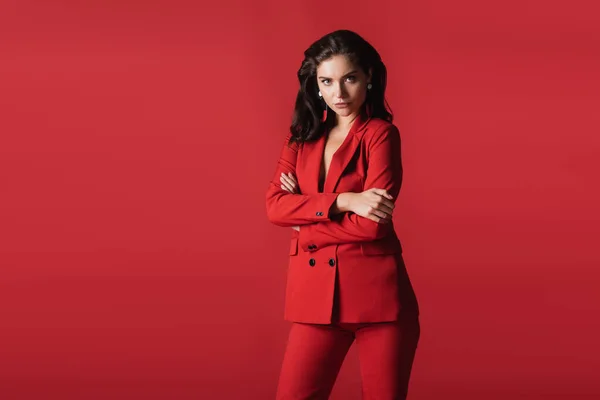 Joven modelo morena en traje posando con los brazos cruzados aislados en rojo - foto de stock