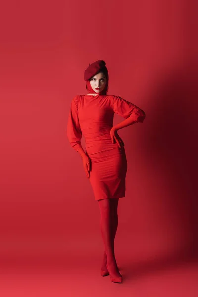 Pleine longueur de jeune femme brune en béret posant avec la main sur la hanche sur le rouge — Photo de stock