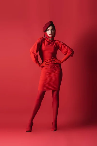 Pleine longueur de jeune femme brune en béret posant avec les mains sur les hanches sur le rouge — Photo de stock