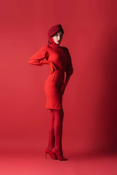 Full length of young brunette woman in beret posing with hand on hip while looking at camera on red — Stock Photo