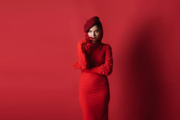 Young brunette woman in beret and dress posing while looking at camera on red — Stock Photo