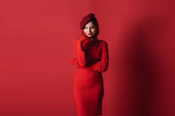 Young pleased woman in beret and dress posing while looking away on red — Stock Photo