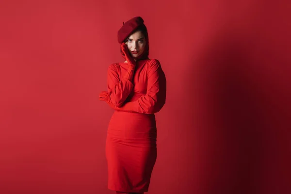 Молода елегантна жінка в береті і плаття позує, дивлячись на камеру на червоному — стокове фото