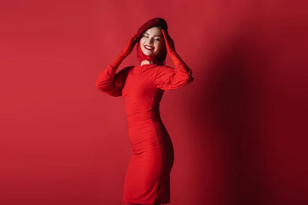 Fröhliche und elegante Frau in Baskenmütze und Kleid posiert auf Rot — Stockfoto