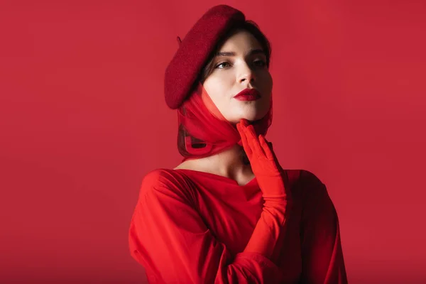 Елегантна молода жінка в береті і рукавичці дивиться вбік і позує ізольовано на червоному — стокове фото