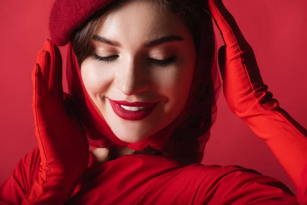 Cheerful young woman adjusting beret isolated on red — Stock Photo