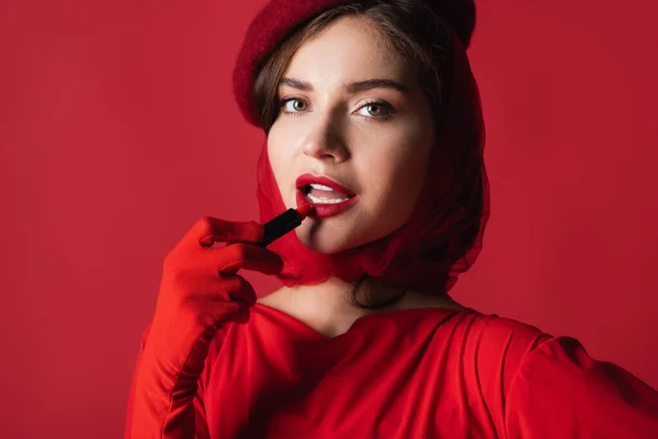 Jovem mulher em luva, lenço de cabeça e boina aplicando batom isolado no vermelho — Fotografia de Stock