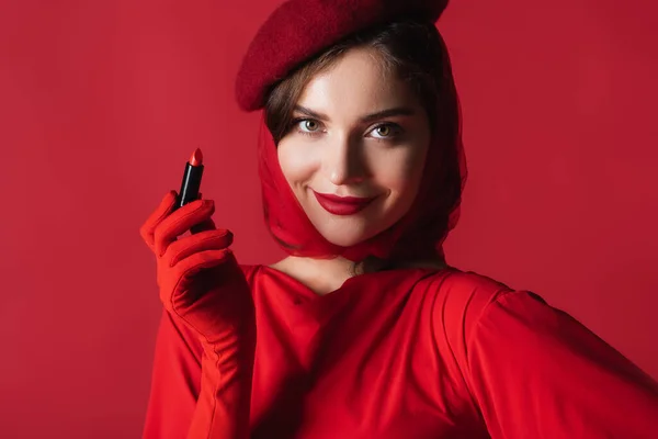 Счастливая женщина в перчатках, платке и берете держа помаду изолированы на красный — стоковое фото
