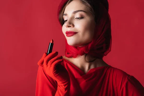 Cheerful woman in glove, headscarf and beret looking at lipstick isolated on red — Stock Photo