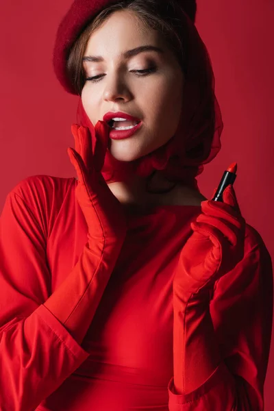 Pretty woman in gloves, headscarf and beret holding lipstick isolated on red — Stock Photo