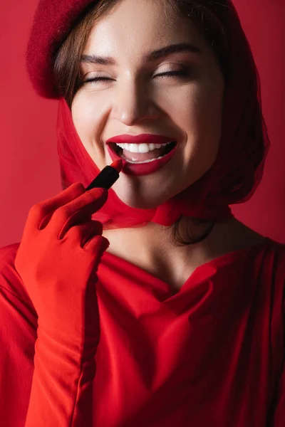 Mulher alegre em luva, lenço de cabeça e boina aplicando batom isolado no vermelho — Fotografia de Stock