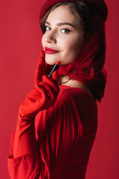 Mujer joven y complacida con guantes, pañuelo para la cabeza y boina con lápiz labial aislado en rojo - foto de stock