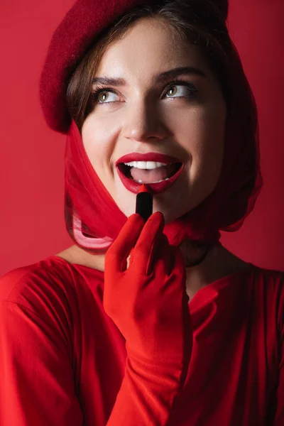 Mujer excitada en guante, pañuelo para la cabeza y boina aplicando lápiz labial aislado en rojo - foto de stock