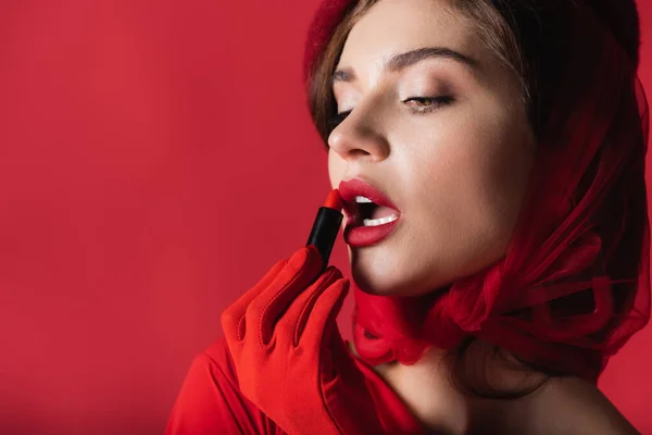 Jolie jeune femme avec bouche ouverte appliquant rouge à lèvres isolé sur rouge — Photo de stock