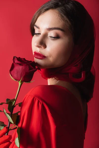 Bonita mujer joven en guante y pañuelo para la cabeza sosteniendo rosa aislado en rojo - foto de stock