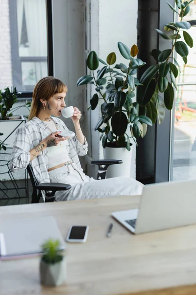 Tätowierte Geschäftsfrau trinkt Kaffee, während sie neben Schreibtisch und Topfpflanze sitzt — Stockfoto