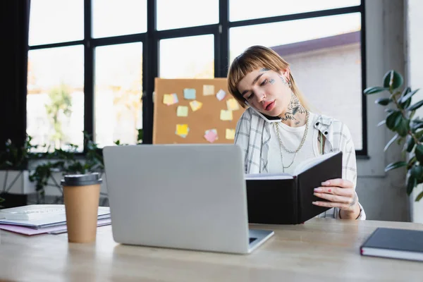 Mulher tatuada sentada na mesa de trabalho com caderno e conversando no celular — Fotografia de Stock