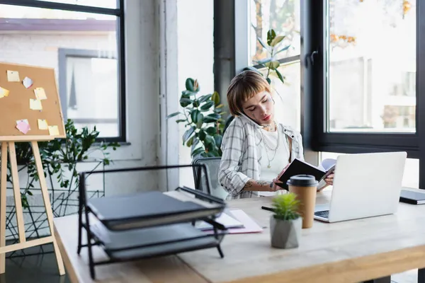 Young tattooed woman with notebook calling on smartphone near laptop in office — Stock Photo