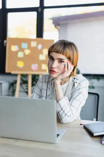 Giovane donna con tatuaggio guardando la fotocamera vicino al computer portatile offuscato in ufficio — Foto stock