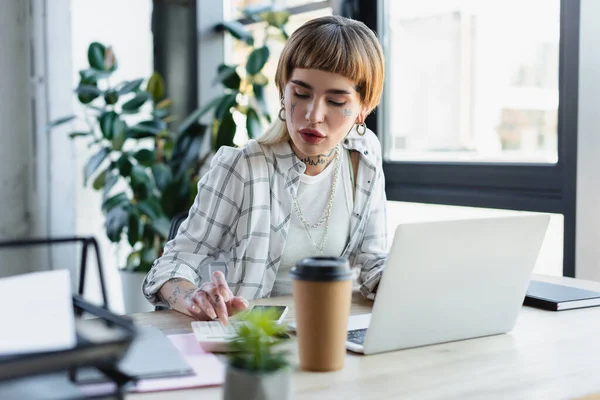 Femme d'affaires à la mode avec corps tatoué en utilisant une calculatrice près d'un ordinateur portable et une tasse en papier floue — Photo de stock