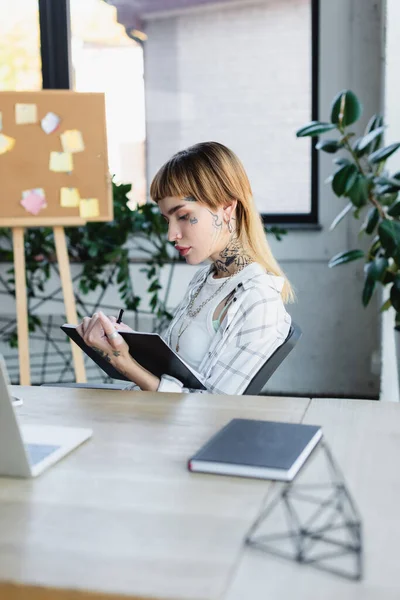 Junge tätowierte Frau sitzt am Arbeitsplatz im Büro und schreibt in Notizbuch — Stockfoto