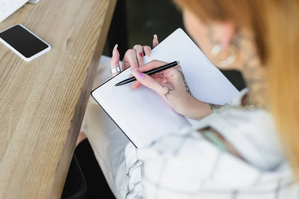 Cropped view of blurred woman with tattoo writing in notebook near smartphone with blank screen on desk — Stock Photo