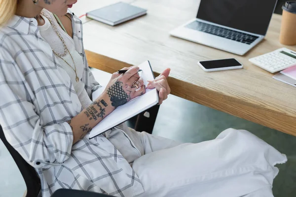 Cropped view of tattooed businesswoman writing in notebook near gadgets with blank screen on desk — Stock Photo