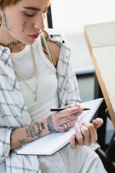 Jovem empresária com corpo tatuado escrevendo no caderno enquanto trabalhava no escritório — Fotografia de Stock