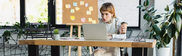 Junge Frau mit Tätowierung hält Pappbecher während der Arbeit am Laptop im Büro, Banner — Stockfoto