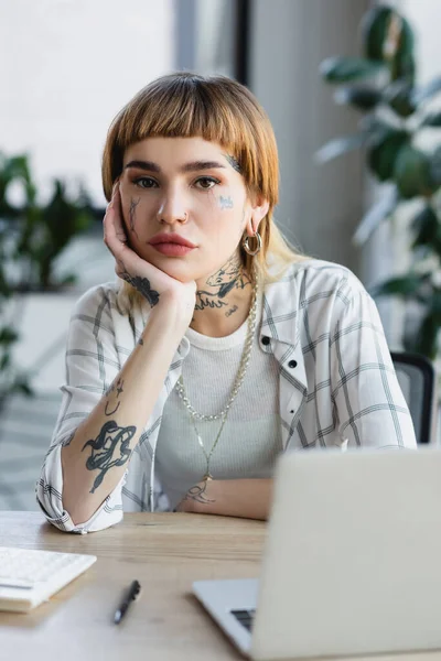 Jeune femme avec tatouage et perçage regardant la caméra tout en étant assis près d'un ordinateur portable flou — Photo de stock