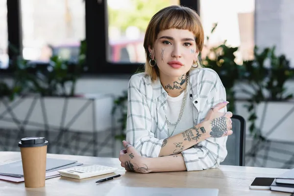 Giovane donna d'affari tatuata seduta con le braccia incrociate sul posto di lavoro in ufficio — Foto stock