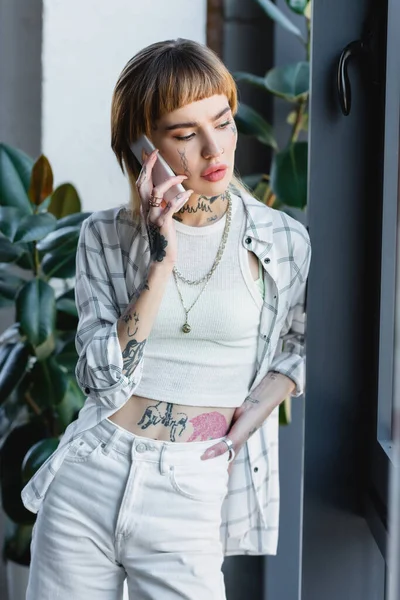 Mulher na moda com o corpo tatuado de pé com a mão no quadril e falando no telefone móvel no escritório — Fotografia de Stock