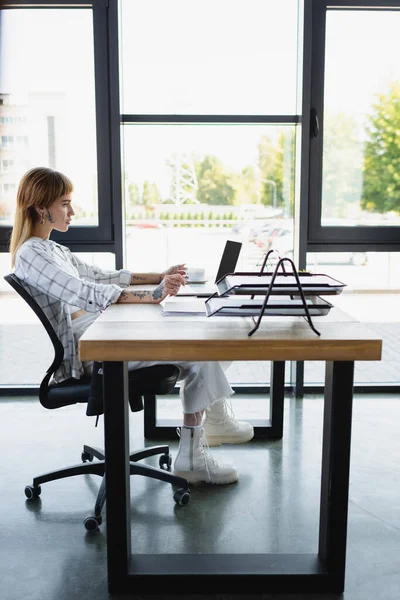 Seitenansicht der jungen und stilvollen Frau am Schreibtisch in der Nähe von Laptop mit leerem Bildschirm — Stockfoto