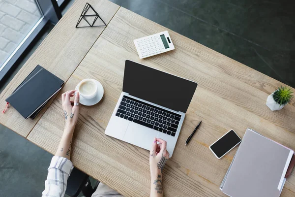 Draufsicht auf beschnittene Frau mit tätowierten Händen in der Nähe von Gadgets mit leerem Bildschirm, Kaffee und Notizbüchern auf dem Schreibtisch — Stockfoto