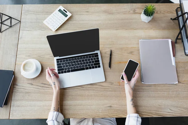 Teilansicht einer Frau mit Tätowierung auf den Händen, die ihr Smartphone in der Nähe eines Laptops mit leerem Bildschirm am Schreibtisch hält — Stockfoto