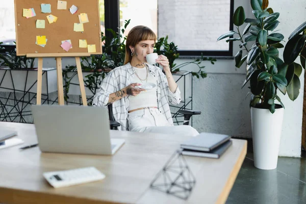 Stylische tätowierte Frau, die Kaffee trinkt, während sie am Schreibtisch neben verschwommenem Laptop sitzt — Stockfoto