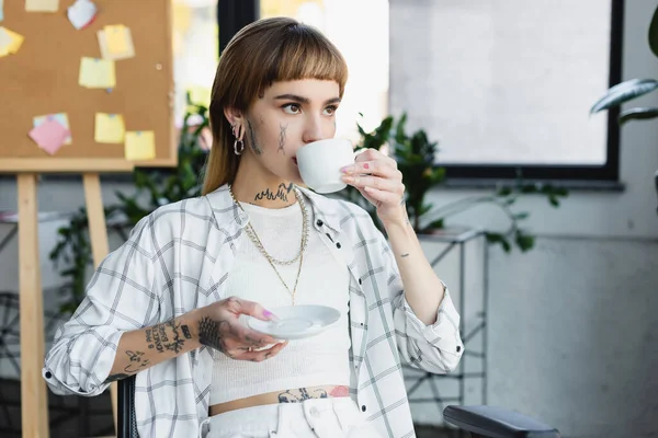 Young tattooed businesswoman with piercing drinking coffee in office — Stock Photo