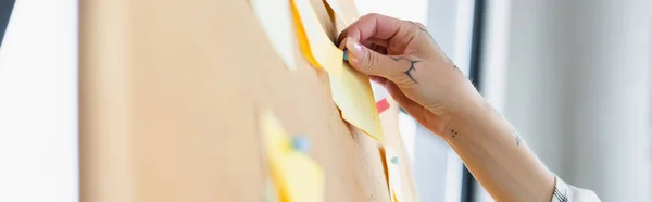 Cropped view of woman with tattooed hand attaching sticky note on corkboard, blurred foreground, banner — Stock Photo