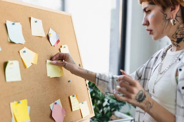 Seitenansicht einer verschwommenen Frau mit Tätowierung, die Notizen auf Korkbrett im Büro anheftet — Stockfoto