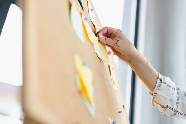 Partial view of woman with tattoo attaching sticky note on blurred corkboard in office — Stock Photo