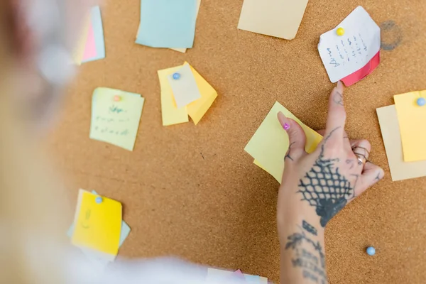 Cropped view of blurred woman with tattoo near corkboard with sticky notes in office — Stock Photo